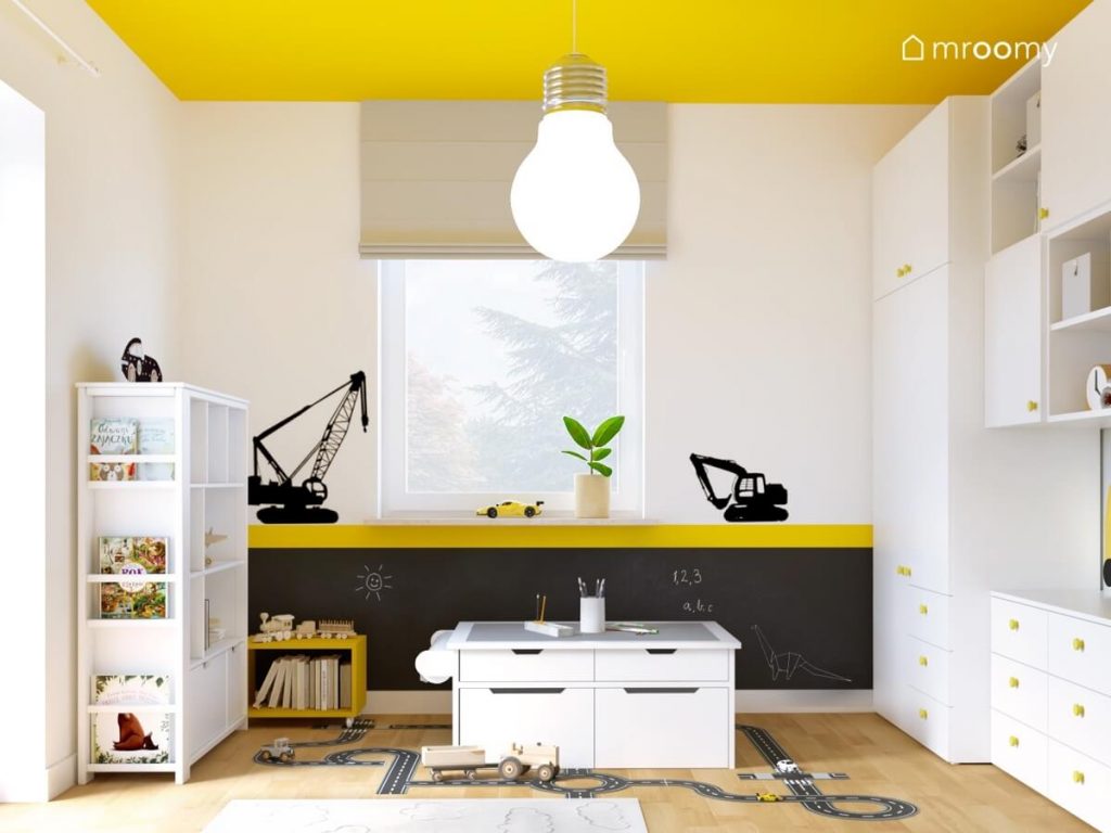 Biało żółty pokój dwulatka a w nim czarna powierzchnia kredowa białe meble stolik do zabawy a na suficie wielka żarówka