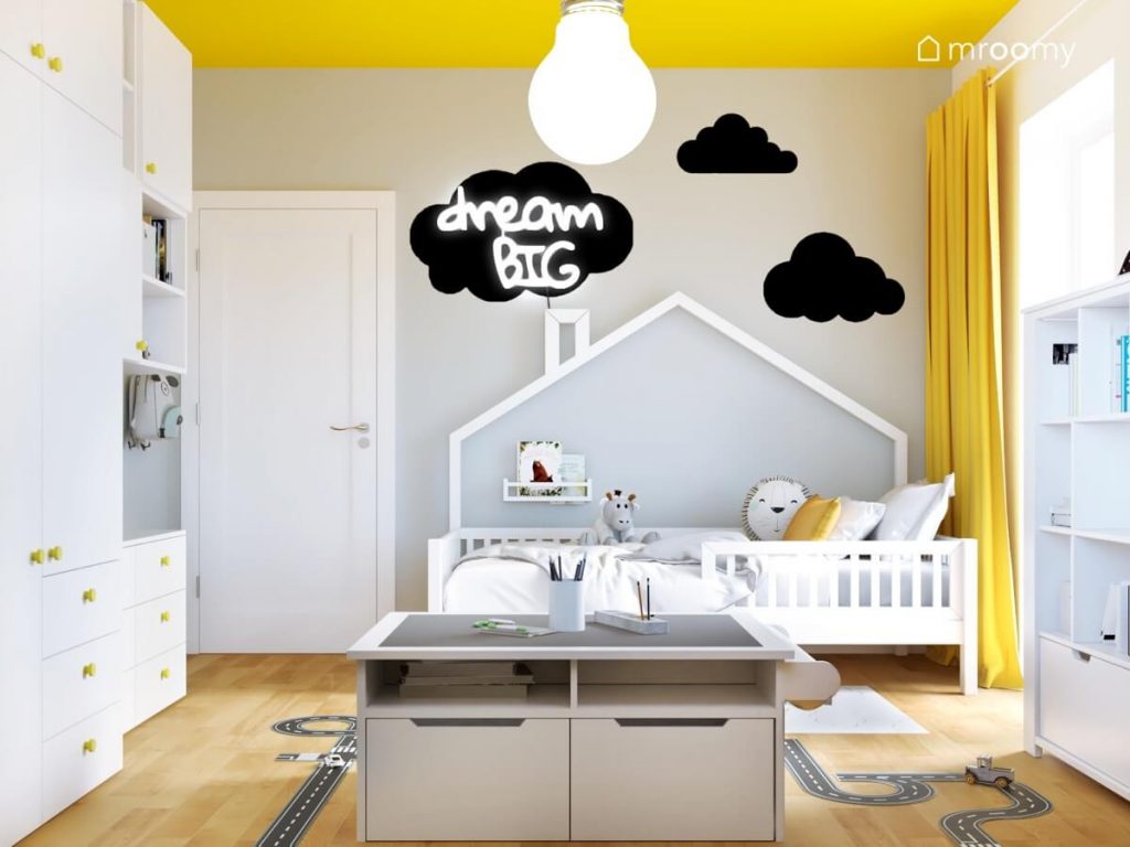 Białe łóżko domek a nad nim trzy czarne chmurki i ledon w kształcie napisu a także stolik do zabawy i białe szafy z żółtymi gałkami w pokoju dla chłopca