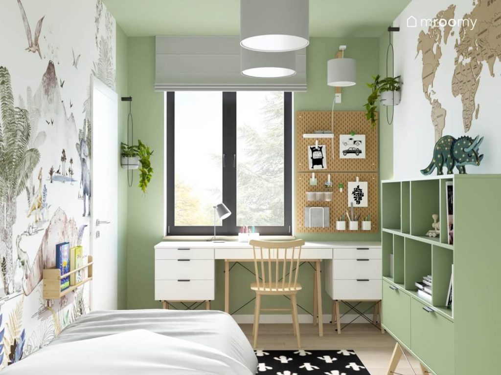 Strefa nauki w biało zielonym pokoju dla chłopca a w niej białe biurko z dwoma kontenerkami a także organizerami ściennymi a nad nimi prosty kinkiet i szara roleta a na ścianach wokół kwietniki