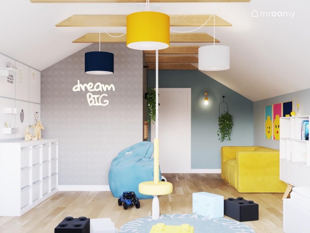 Biało szaro niebieski poddaszowy pokój dla chłopca a w nim biały regał z pojemnikami żółta kanapa żółta huśtawka ring błękitna pufa pufki klocki oraz ściana pokryta tapetą we wzór klocka Lego i ledon w kształcie napisu i kwietniki