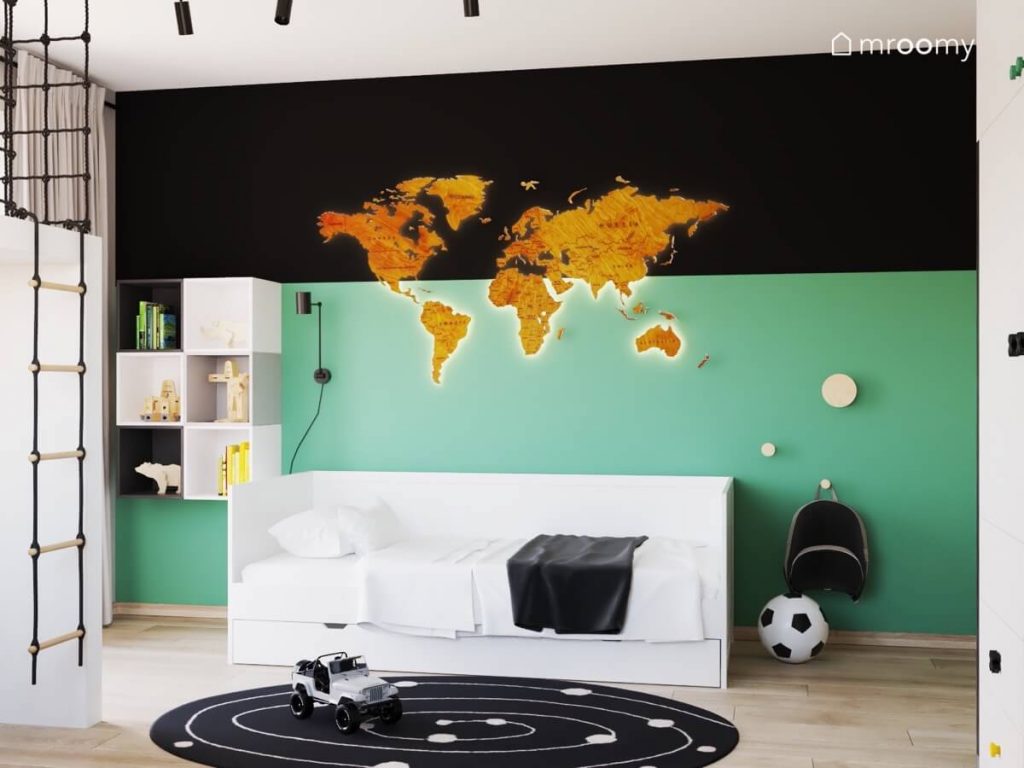 Podświetlana drewniana mapa świata na czarno zielonej ścianie w pokoju chłopca a oprócz tego białe łóżko kompozycja szafek ściennych i wieszaki ze sklejki