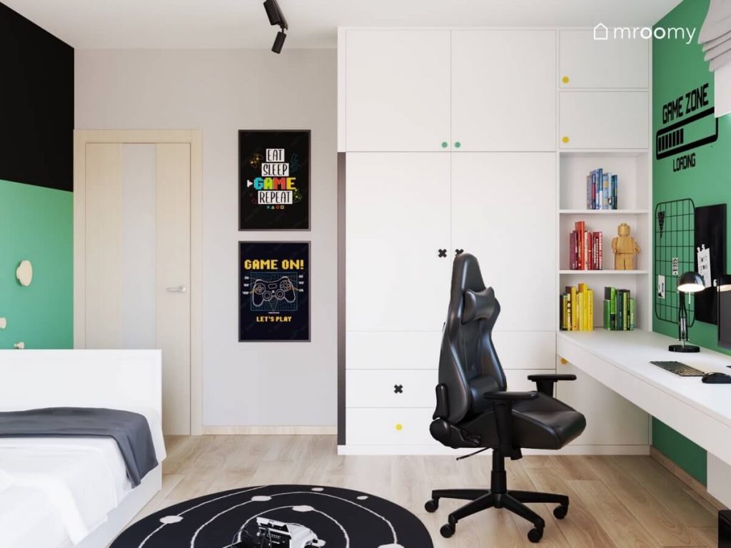 Biało szaro czarno zielony pokój chłopca w wieku szkolnym a w nim białe biurko z fotelem gamingowym biała szafa z kolorowymi gałkami oraz plakatami gamingowymi