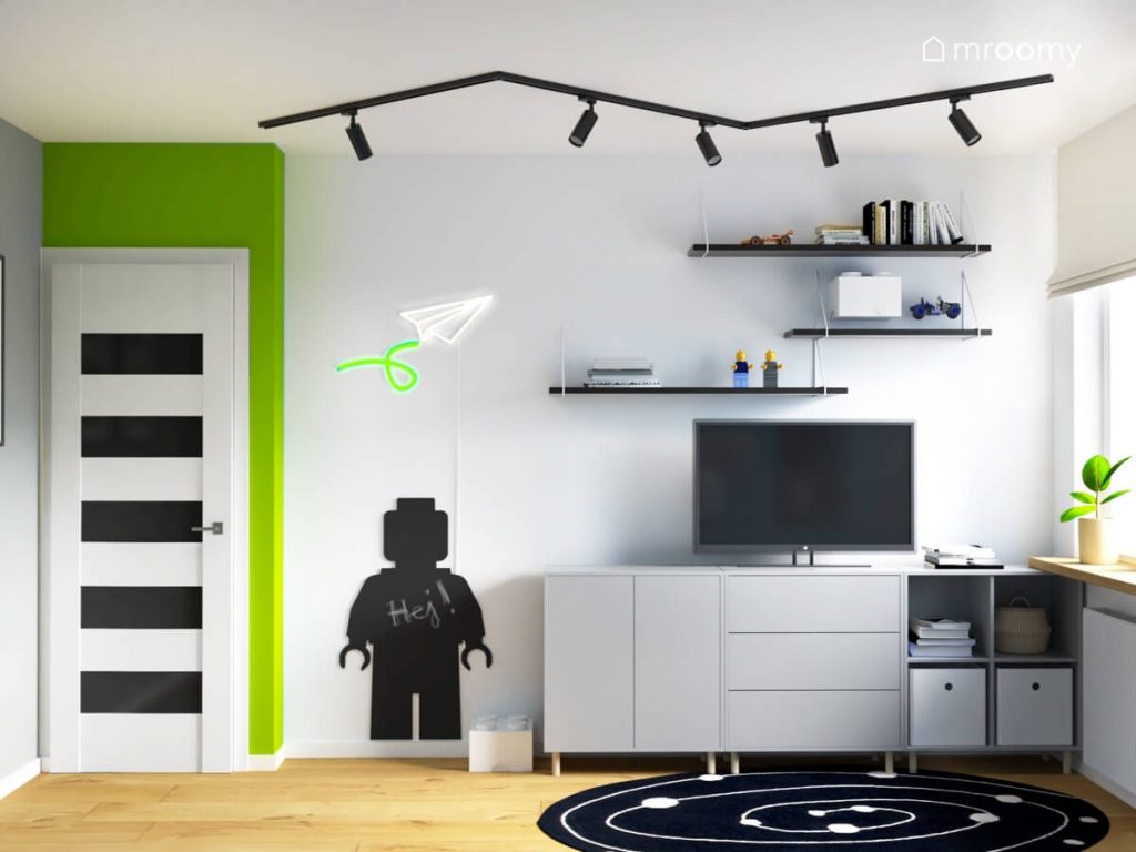 Biała ściana w pokoju dla chłopca a na niej tablica kredowa w kształcie ludzika Lego ledon w kształcie papierowego samolotu półki oraz szafka pod telewizor a na suficie reflektory na szynie