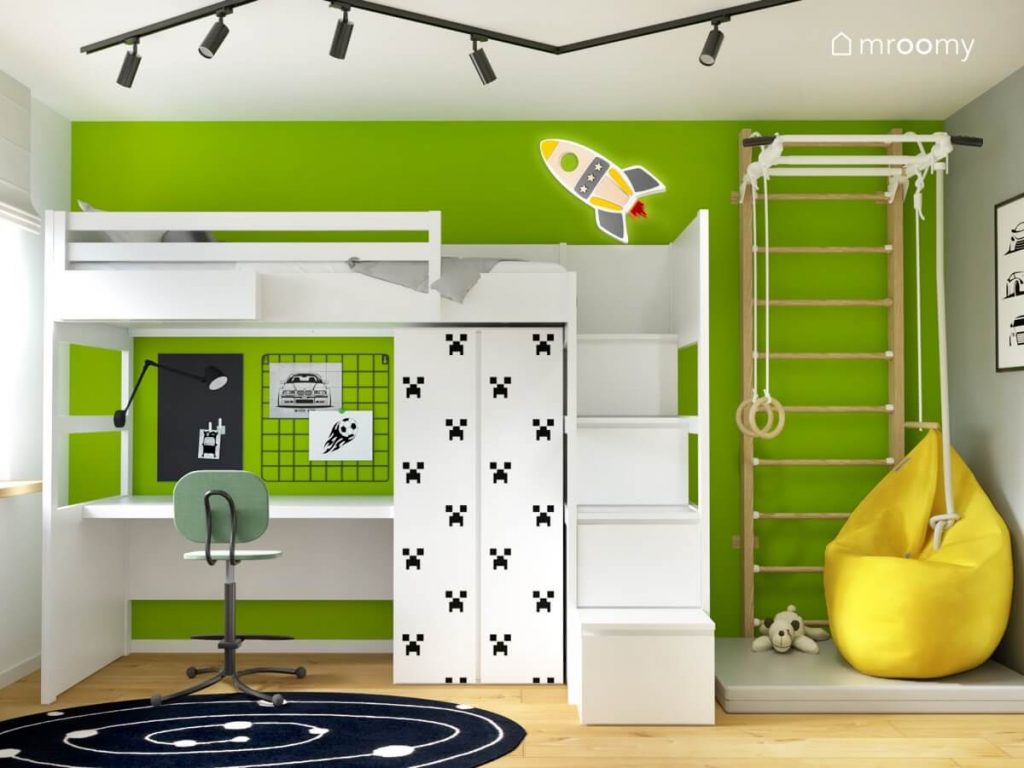 Soczyście zielona ściana w pokoju dla chłopca w wieku szkolnym a na niej biała antresola z biurkiem pod spodem szafa z motywami Minecraft oraz drabinka gimnastyczna i lampka w kształcie rakiety