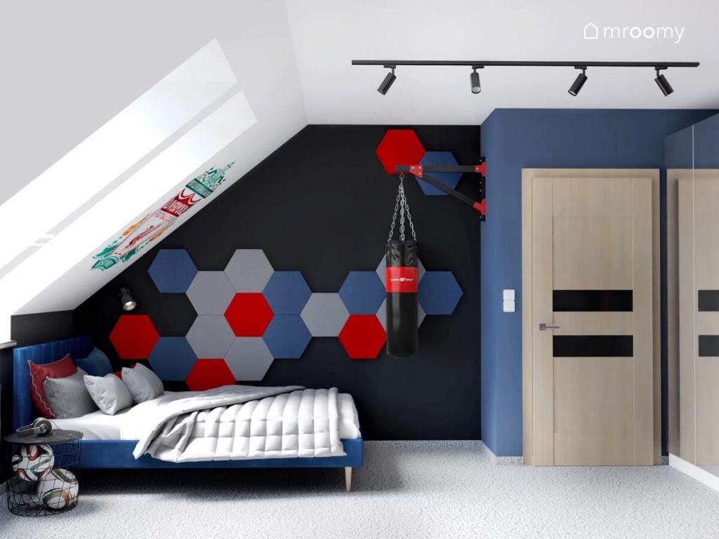 Strefa spania w poddaszowym pokoju chłopca a w niej niebieskie tapicerowane łóżko a nad nim kompozycja z sześciokątnych paneli ściennych w trzech kolorach a obok worek treningowy