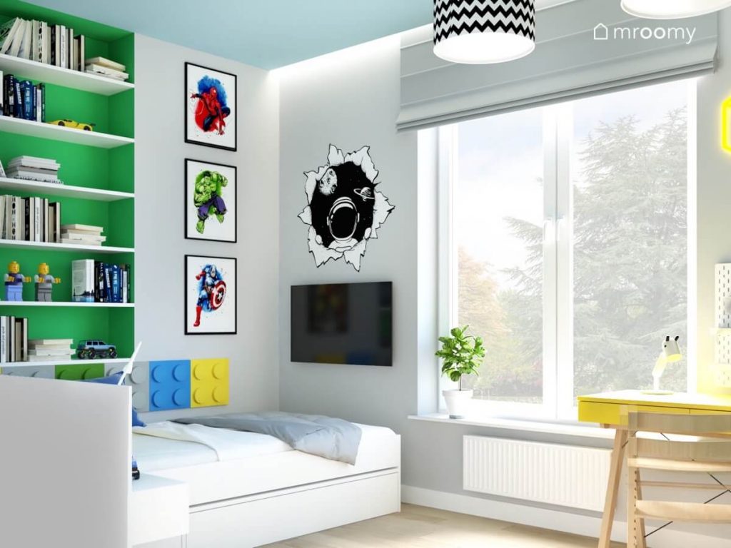 Jasny pokój dla kilkuletniego chłopca a w nim białe łóżko uzupełnione panelami w kształcie klocków telewizor zielona wnęka z półkami plakaty z superbohaterami oraz kosmiczna naklejka