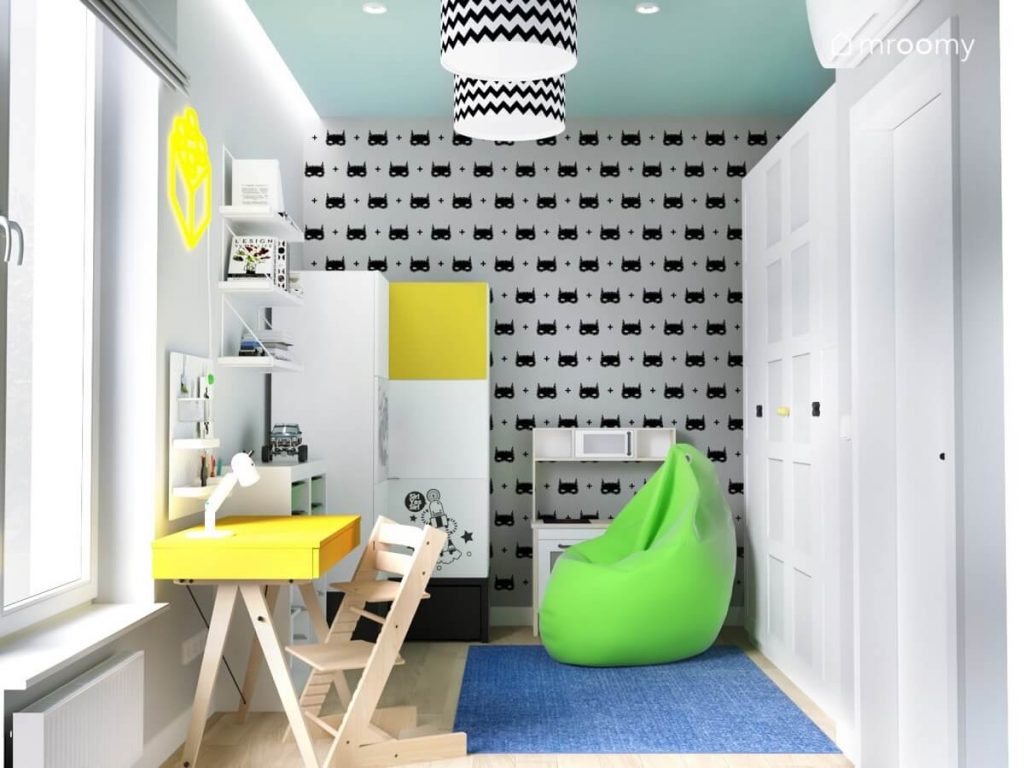 Ściana w pokoju chłopca pokryta tapetą w Batmany a także duża biała szafa mniejsza szafa z nakładkami na fronty soczyście zielona pufa niebieski dywan żółte biurko oraz kuchnia dla dzieci