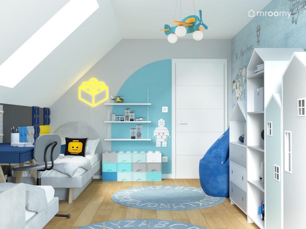 Biało szaro błękitny pokój dwóch chłopców a w nim regały w kształcie domków liczne motywy Lego oraz lampa samolot