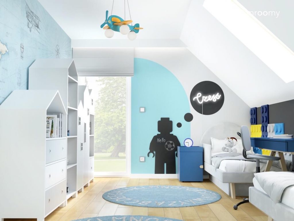 Jasny poddaszowy pokój dwóch chłopców a w nim szare tapicerowane łóżka z biurkiem pomiędzy meble w kształcie domków oraz tablica kredowa w kształcie ludzika Lego