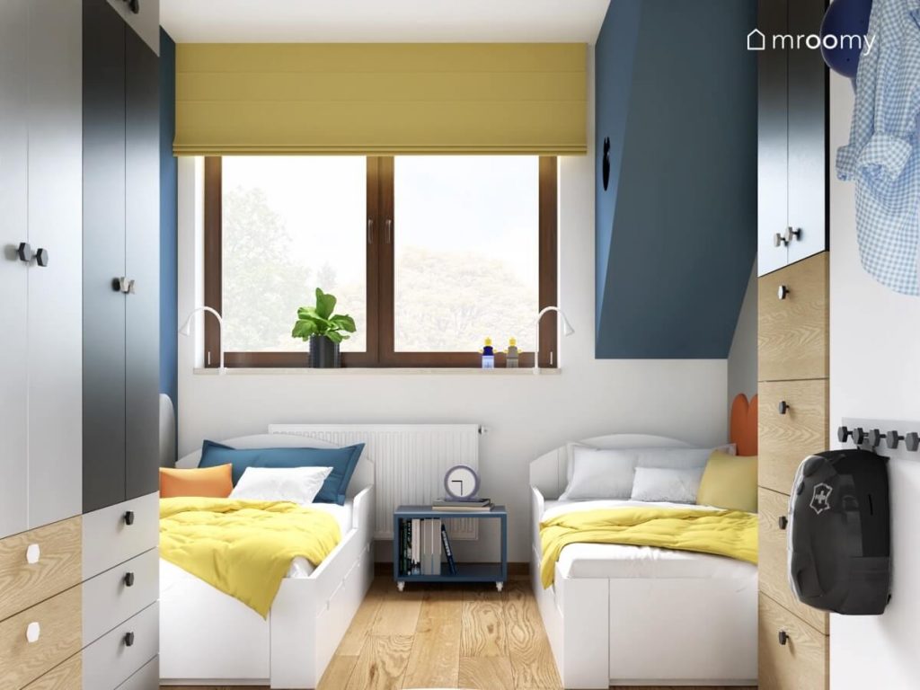 Przytulna sypialnia dwóch chłopców a w niej dwa białe łóżka niebieski stolik nocny na kółkach oraz żółta roleta