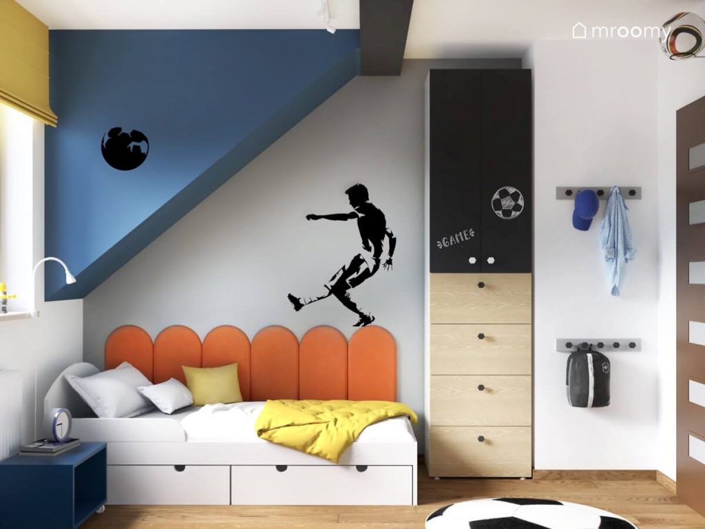 Białe łóżko z szufladami a na ścianie pomarańczowe panele ścienne i naklejka w kształcie piłkarza a obok wąska szafa z drewnianymi szufladami i powierzchnią kredową w sypialni dwóch braci