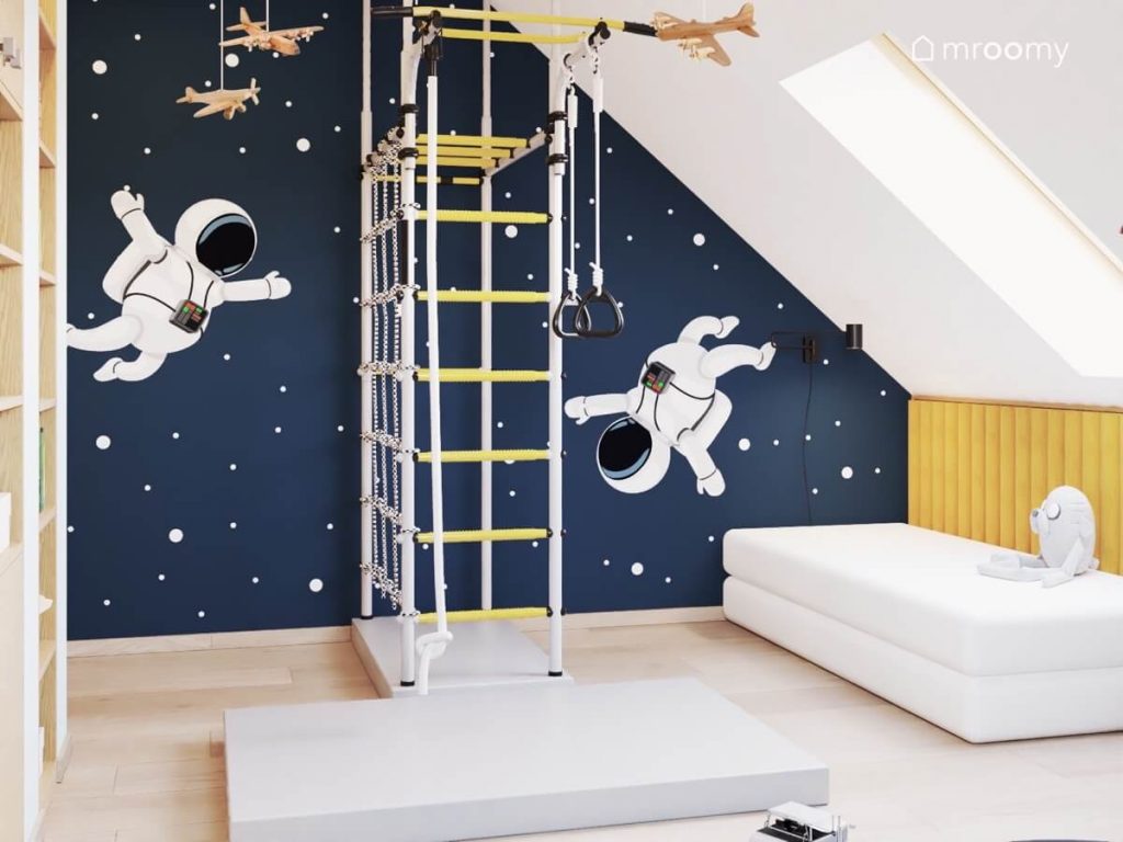 Kosmiczna granatowa ściana z naklejkami w kształcie astronautów a na niej rozbudowana drabinka gimnastyczna a u sufitu drewniane samoloty w poddaszowym pokoju dwóch chłopców