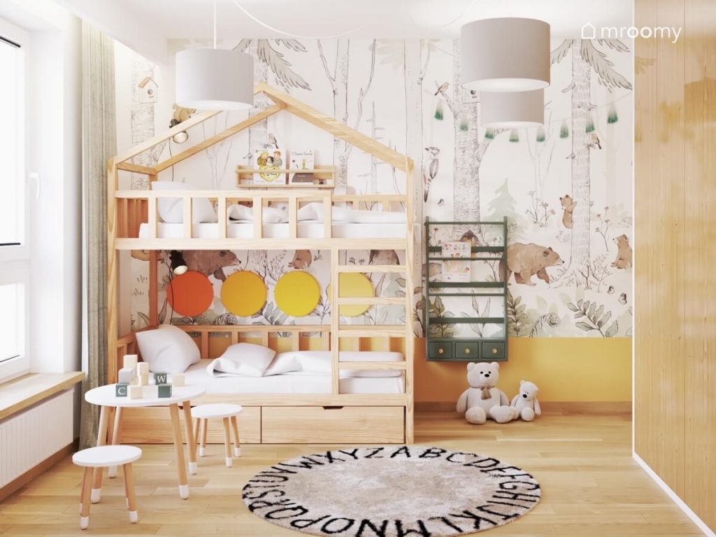 Drewniane łóżko piętrowe w kształcie domku a obok zielona biblioteczka a na ścianie leśna tapeta i girlanda pomponów a na podłodze dywan z alfabetem w pokoju dwóch chłopców