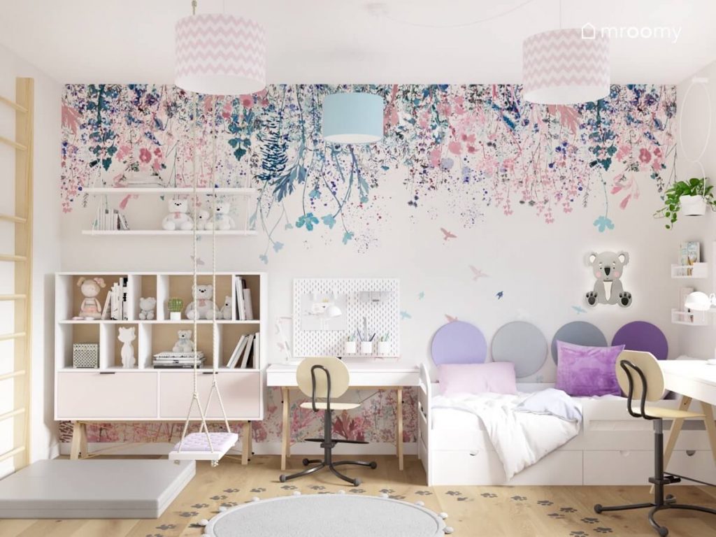 Biały regał z różowymi szufladami huśtawka pokojowa białe półki ścienne biurko z organizerami ściennymi oraz białe łóżko a na ścianie tapeta w kwiaty w pokoju dla dwóch dziewczynek