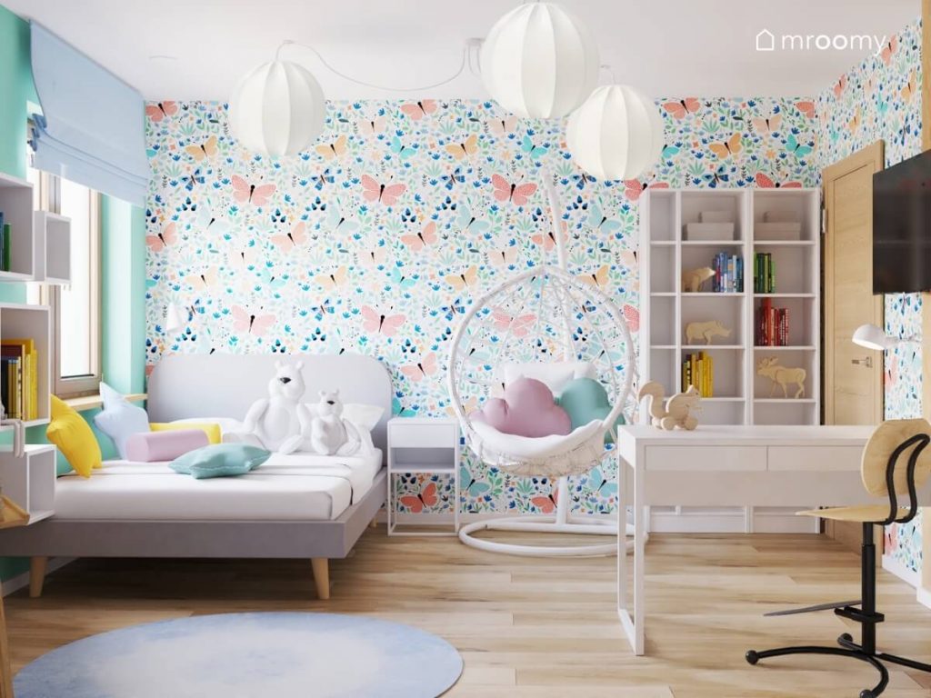 Biało niebieski pokój dla dziewczynki a w nim szare łóżko białe meble fotel wiszący lampa sufitowa z papierowymi kloszami a na ścianie radosna tapeta w motyle