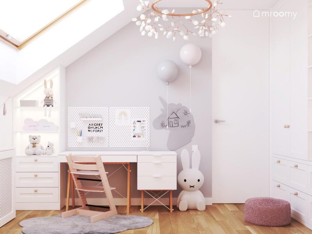 Białe biurko na drewnianych nogach z kontenerkiem a na ścianie organizery tablica kredowa w kształcie jednorożca kinkiety w kształcie balonów a na podłodze lampa królik w pokoju małej dziewczynki