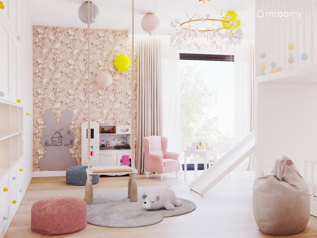 Jasny pokój dla dziewczynki a w nim białe meble huśtawka pokojowa dywan w kształcie chmurki niskie pufy tablica kredowa w kształcie królika oraz tapeta w wiewiórki na ścianie