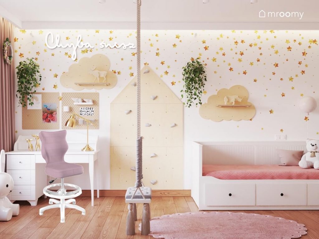 Biała ściana w złote gwiazdki w pokoju dziewczynki a na niej ścianka wspinaczkowa w kształcie domku półki w kształcie chmurek kwietnik ledon w kształcie napisu oraz biurko z organizerami