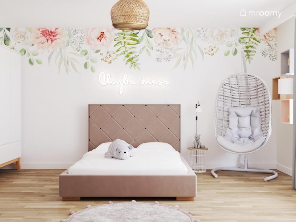 Duże tapicerowane łóżko w jasnym pokoju dziewczynki a na ścianie za nim ledon w kształcie napisu oraz kwiaty a obok fotel wiszący