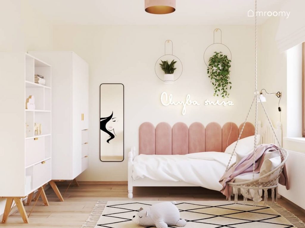 Jasny pokój dla nastolatki a w nim białe meble na drewnianych nogach podłużne lustro łóżko uzupełnione różowymi panelami a nad nim ledon w kształcie napisu oraz kwietniki