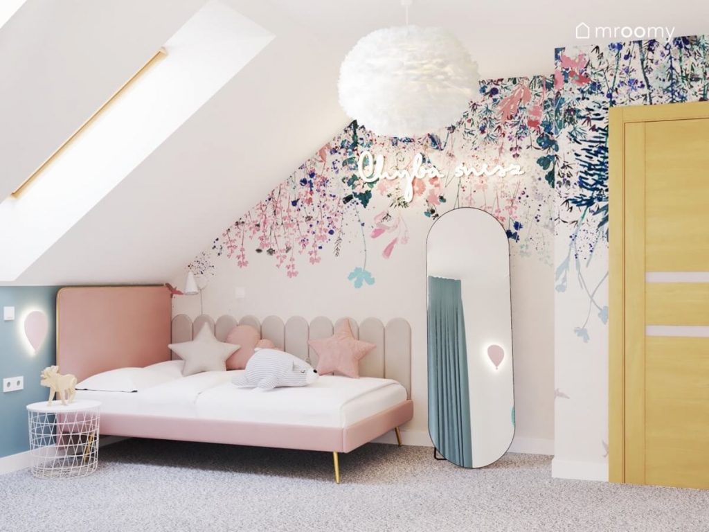 Różowe łóżko z wysokim zagłówkiem i szarymi panelami ściennymi na boku a obok podłużne lustro a na ścianie tapeta w kwiaty i ledon w kształcie napisu w pokoju dziewczynki