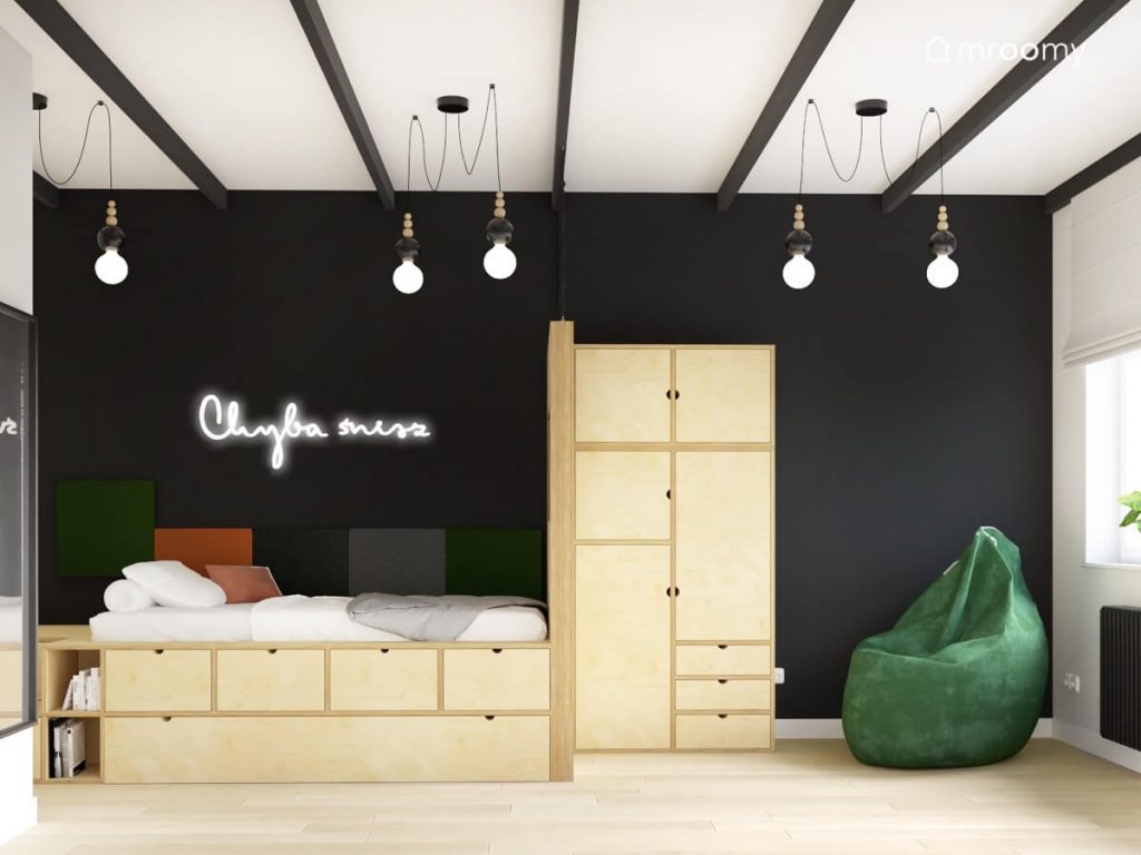 Czarna ściana w pokoju dla nastolatka a na niej ledon w kształcie napisu oraz panele ścienne w ciemnych kolorach a także drewniane łóżko i szafa oraz ciemnozielona pufa a na suficie ozdobne lampy