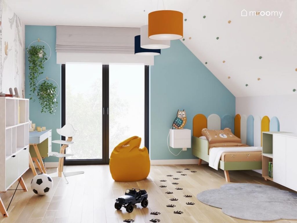 Biało niebieski poddaszowy pokój dla chłopca a w nim zielone łóżko biały regał na drewnianych nogach oraz pomarańczowe dodatki a na podłodze psie łapki