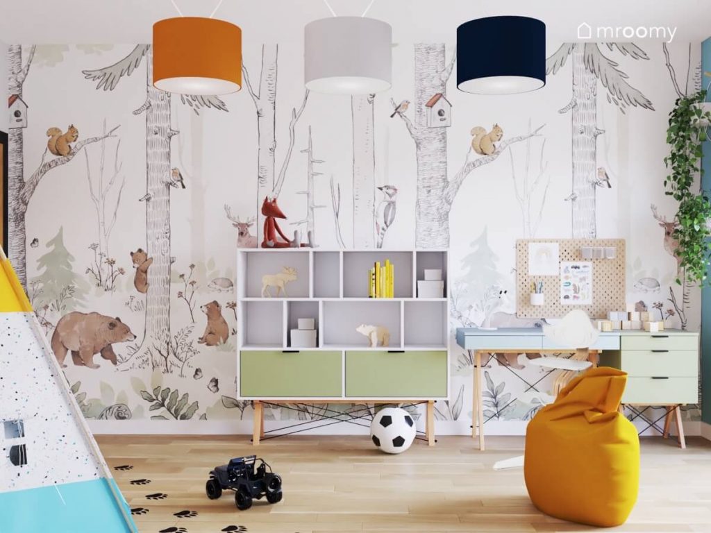 Ściana w pokoju małego chłopca pokryta leśną tapetą a na niej biały regał z zielonymi szufladami niebieskie biurko na drewnianych nogach i zielony kontenerek a na suficie pomarańczowa szara i granatowa lampa