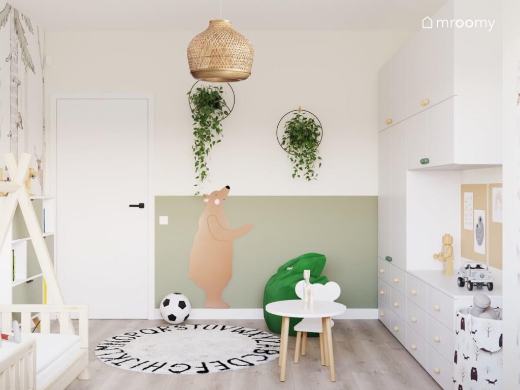 Biało zielona ściana w pokoju dla małego chłopca a na niej dwa ozdobne kwietniki oraz naklejka z niedźwiedziem a na podłodze zielona pufa dywan z alfabetem oraz mały stolik z krzesełkiem z oparciem w kształcie misia