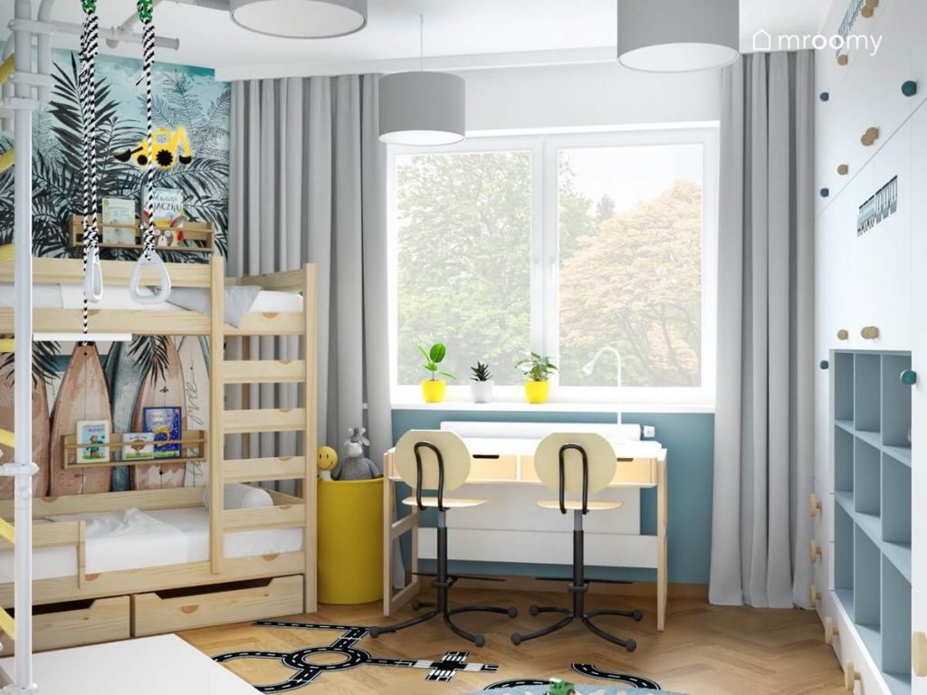 Drewniane łóżko piętrowe z półeczkami na książki biało drewniane biurko z krzesłami z podnóżkami a także żółty worek na zabawki szare zasłony i naklejka podłogowa w kształcie jezdni w pokoju dwóch chłopców