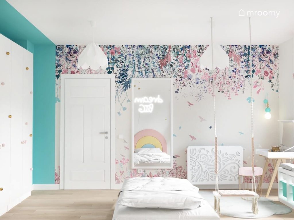Ściana pokryta kwiecistą tapetą a na niej prostokątne lustro a na suficie lampy bezy w pokoju dziewczynki