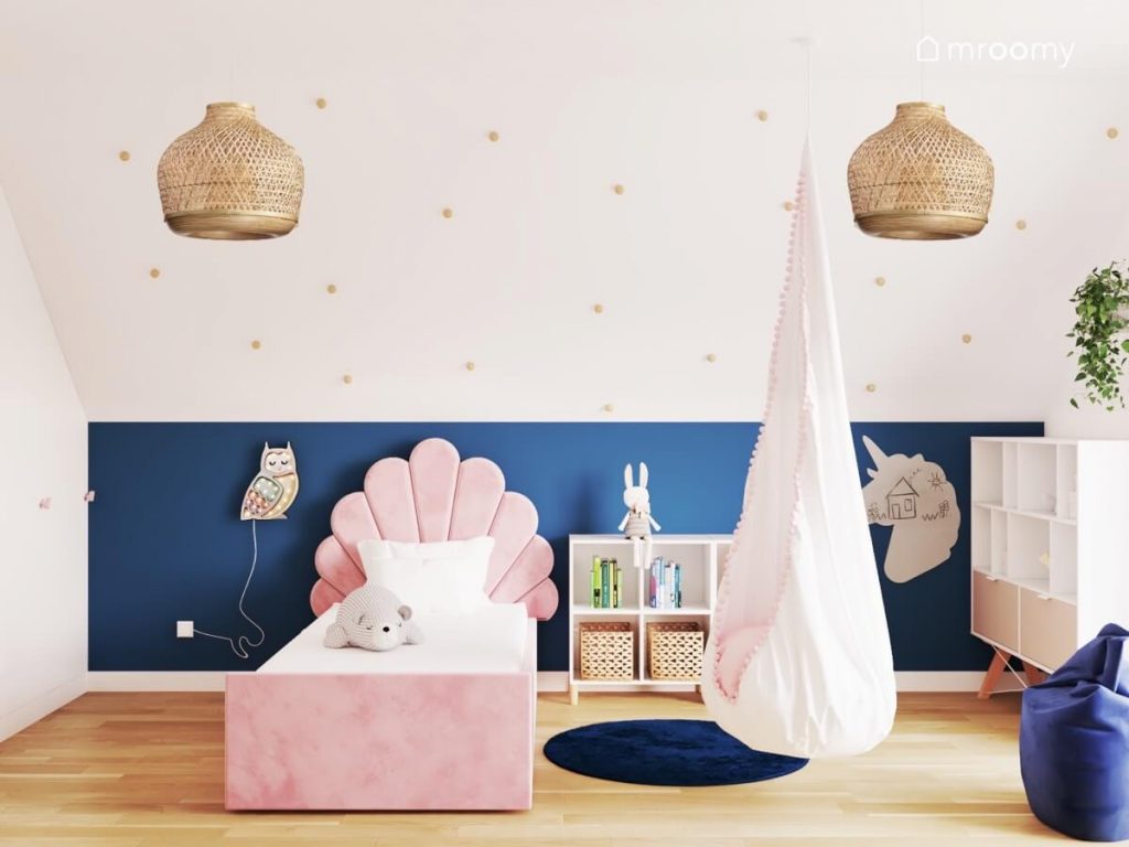 Biało granatowy pokój dziewczynki a w nim skos ozdobiony gałkami różowe łóżko z ozdobnym oparciem fotel kokon tablica kredowa w kształcie jednorożca lampka sówka i lampy sufitowe w bambusowych kloszach
