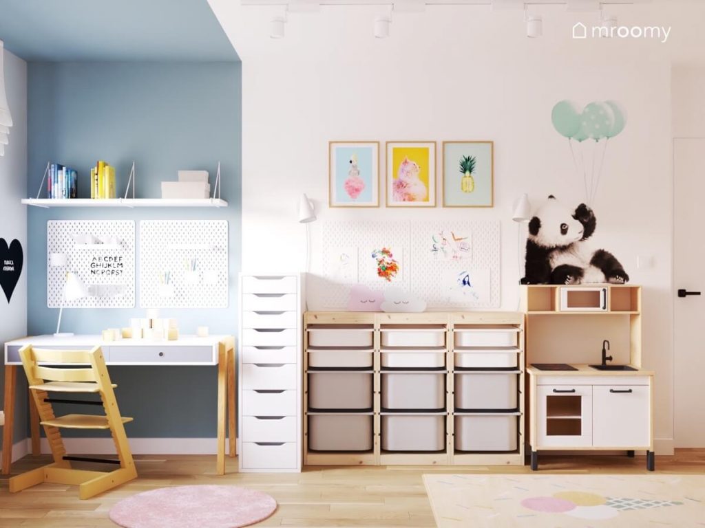 Niebieska strefa nauki w pokoju dla dziewczynki a w niej białe biurko półki i organizery a obok regał z pojemnikami ozdobne plakaty naklejka z pandą i kuchenka dla dzieci