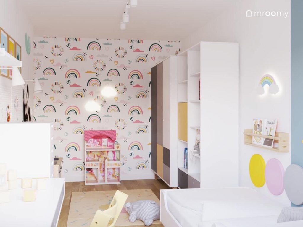Ściana pokryta kolorową tapetą w tęcze a na niej domek do zabawy i lampki w kształcie chmurek a obok białe meble z różnokolorowymi frontami w pokoju dziewczynki