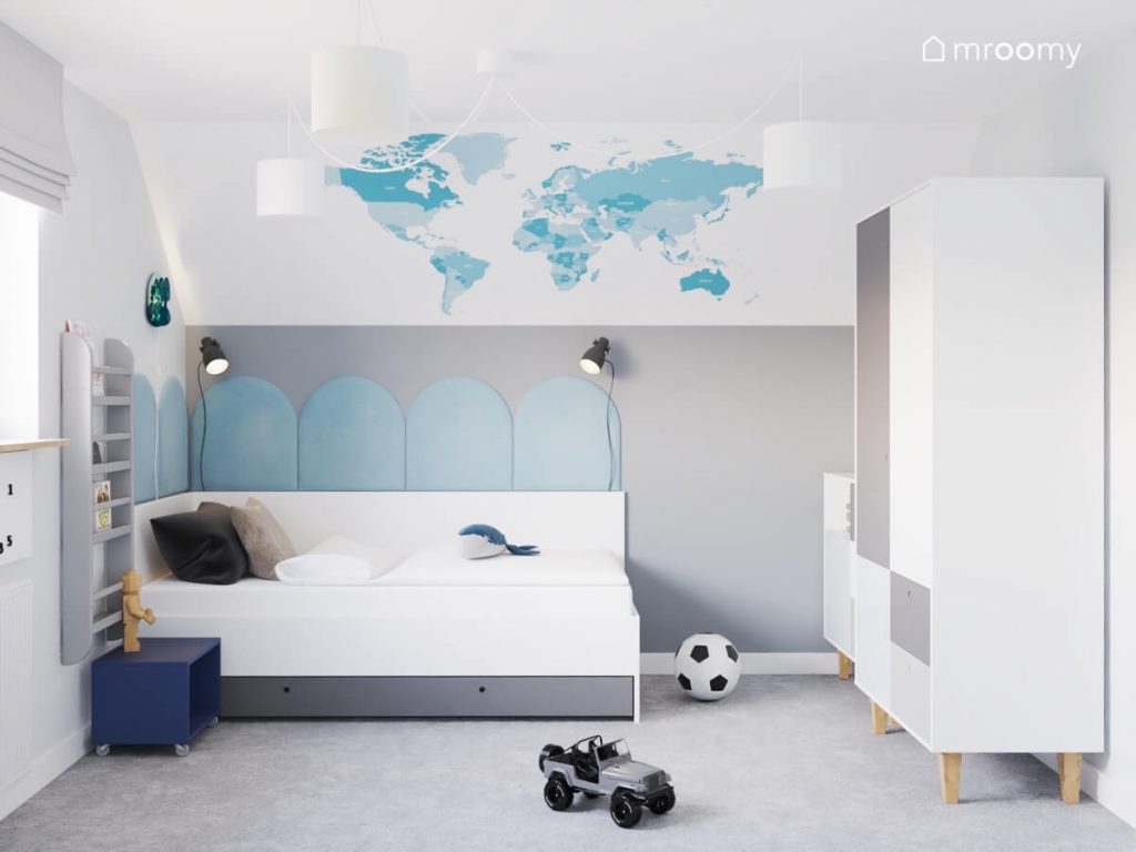 Biało szare łóżko uzupełnione błękitnymi panelami a obok granatowy stolik nocny na kółkach a na ścianie błękitna mapa świata w biało szarym pokoju chłopca