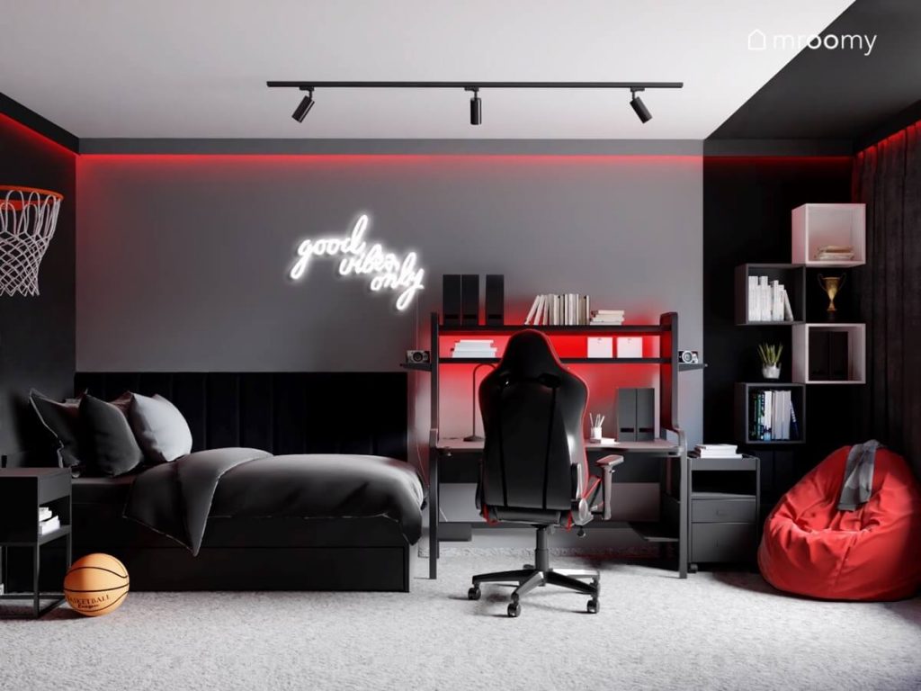 Biało szaro czarny pokój nastolatka a w nim czarne łóżko czarne panele ścienne podświetlane na czerwono biurko szafki ścienne ledon w kształcie napisu oraz czerwony worek do siedzenia