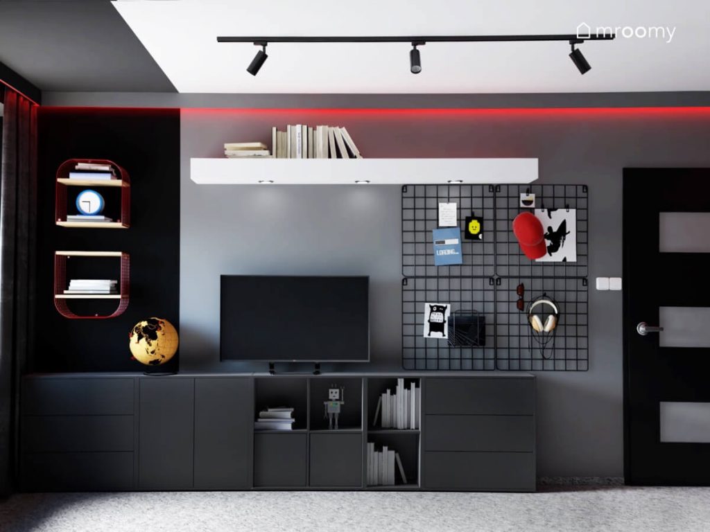 Niskie czarne meble a na nich telewizor a na ścianie organizery oraz półki oraz biała półka na książki w szaro czarno białym pokoju dla nastolatka z czerwonymi akcentami