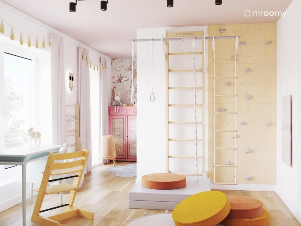Drewniana drabinka gimnastyczna oraz ścianka wspinaczkowa ze sklejki a na podłodze żółte i pomarańczowe pufy w pokoju dziewczynki