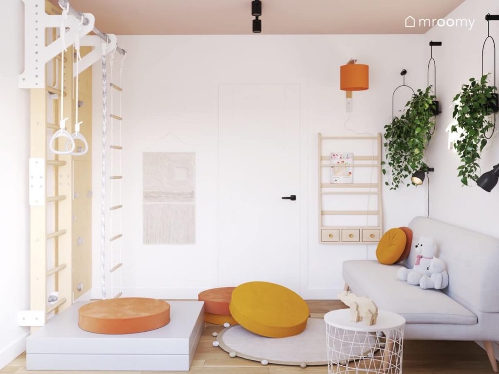 Biały pokój dziewczynki w wieku przedszkolnym z beżowym sufitem a w nim drabinka gimnastyczna ścianka wspinaczkowa jasnoszara sofa drewniana biblioteczka oraz kwietniki i pomarańczowe dodatki