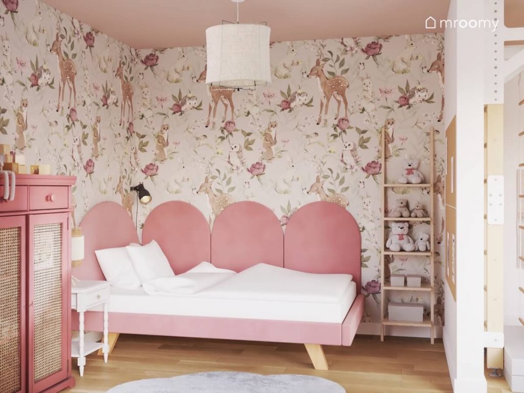 Strefa spania w pokoju małej dziewczynki a w niej różowe tapicerowane łóżko uzupełnione z boku panelami ściennymi w podobnym kolorze a obok regał a na ścianie tapeta w zwierzęta i kwiaty