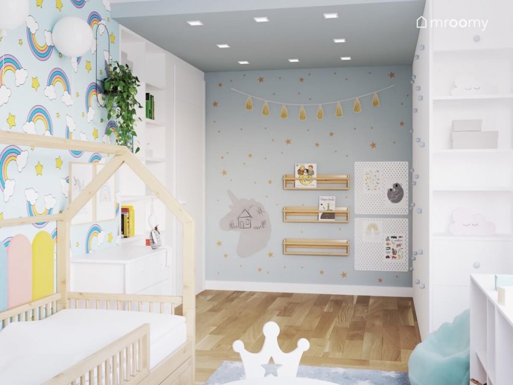 Biało szarobłękitny pokój dziewczynki a w nim złote gwiazdki i girlanda pomponów na ścianie a także półeczki drewniane organizery i tablica kredowa w kształcie jednorożca