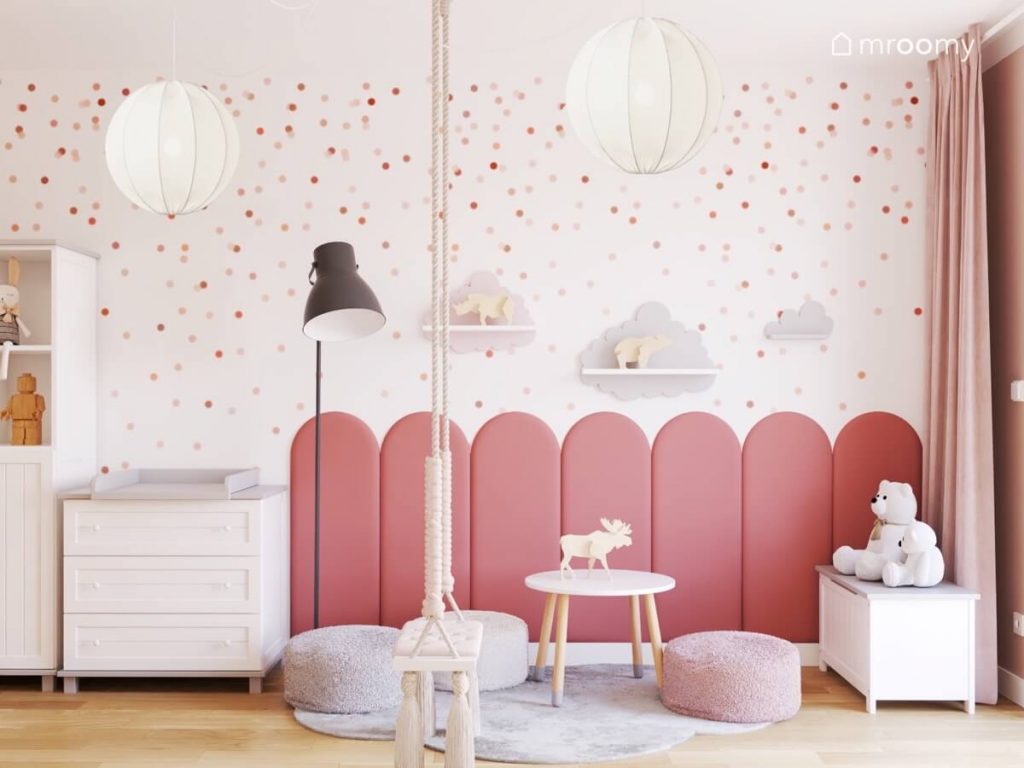 Kącik do zabawy w pokoju dziewczynki uzupełniony różowymi panelami ściennymi a w nim mały stolik trzy pufy dywan w kształcie chmurki skrzynka z siedziskiem lampa oraz huśtawka wisząca