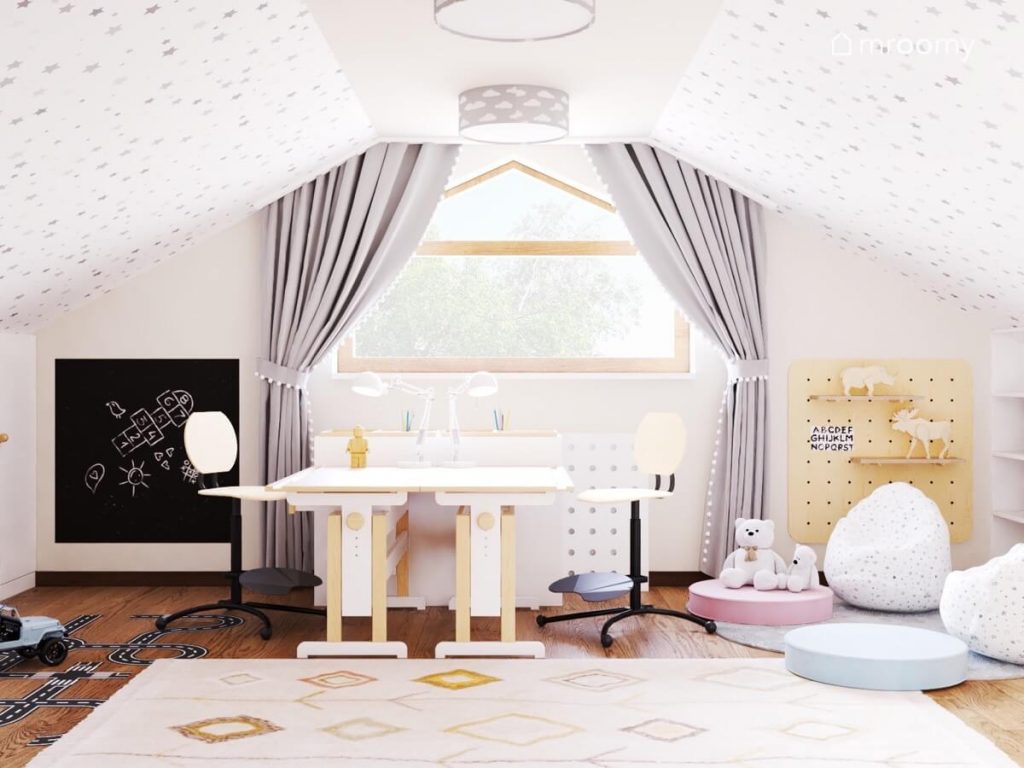Poddaszowy pokój dla chłopca i dziewczynki a w nim biało drewniane biurka czarna tablica kredowa szare zasłony niskie pufki oraz wzorzysty dywan