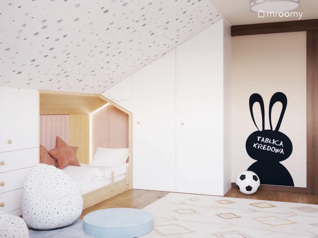 Drewniane łóżko dziewczynki uzupełnione różowymi panelami ściennymi a także biała szafa i tablica kredowa w kształcie królika a na suficie gwiazdki w poddaszowym pokoju rodzeństwa