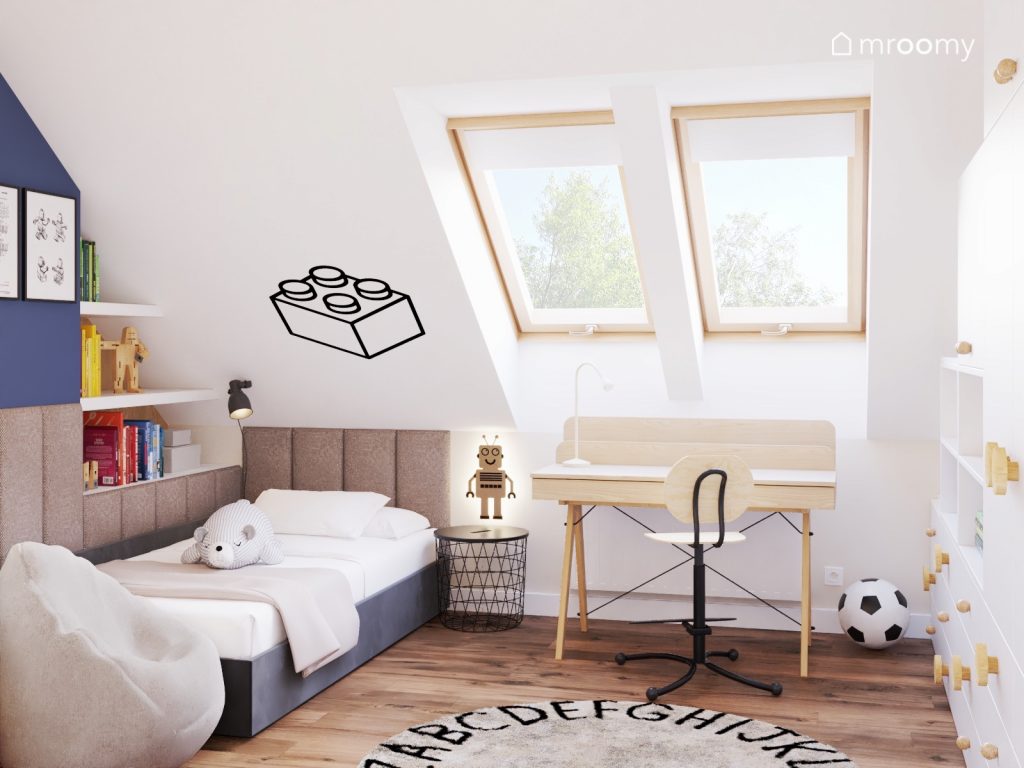 Jasny poddaszowy pokój dla chłopca z czarnymi łóżkiem uzupełnionym miękkimi panelami drewnianym biurkiem oraz naklejką w kształcie klocka i dywanem z alfabetem