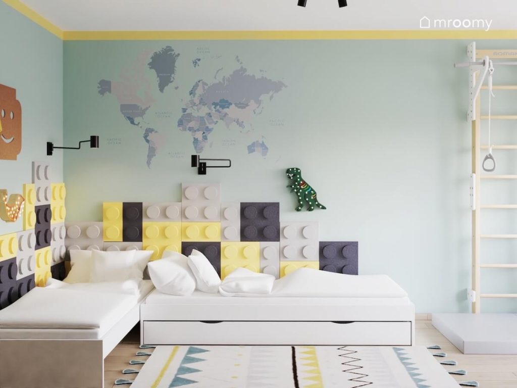 Dwa jasne łóżka w pokoju dwóch braci a za nimi zielono żółta ściana uzupełniona panelami w kształcie klocków oraz mapą świata