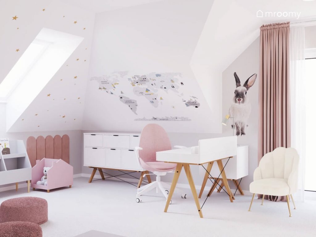 Biała komoda na drewnianych nogach oraz biurko z tej samej kolekcji z różowym krzesłem a na ścianach naklejki w kształcie królika mapy świata i złotych gwiazdek w pokoju dziewczynki