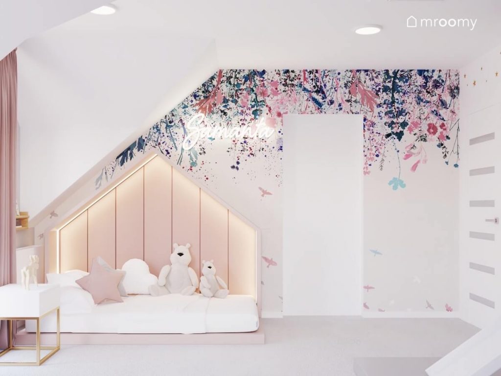 Trójkątne łóżko wyłożone miękkimi panelami a za nim tapeta w kwiaty i ledon w kształcie imienia w pokoju siedmiolatki
