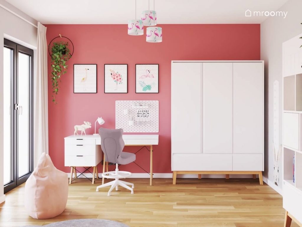 Ciemnoróżowa ściana w pokoju dziewczynki a na niej ozdobne plakaty z flamingami kwietnik oraz organizer a także duża biała szafa oraaz biurko z kontenerkiem