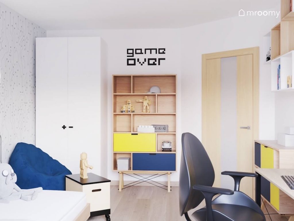 Wysoka biała szafa a w obok drewniany regał z żółtą i granatową szufladą a nad nim gamingowa naklejka w pokoju fana gier komputerowych